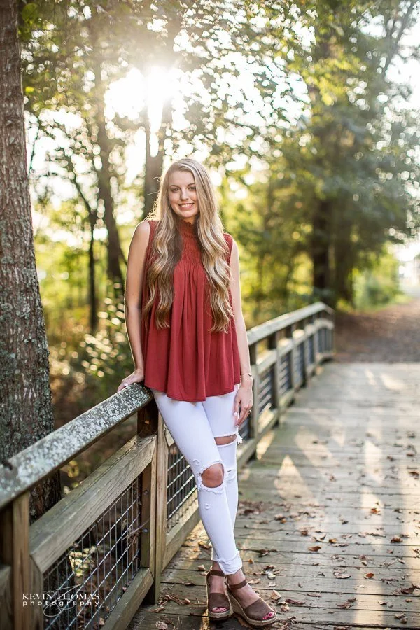 A young woman with long blonde hair wearing a sleeveless red top, white ripped jeans, and brown wedge sandals standing on a wooden pathway in a wooded park, smiling at the camera with sunlight filtering through the trees.