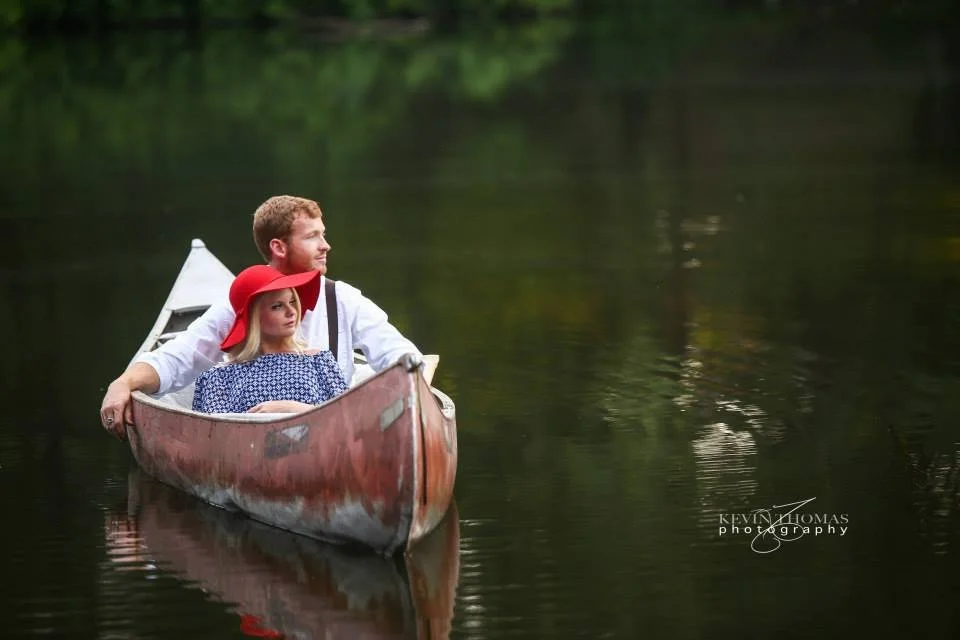 A man and a woman in a canoe on a calm river, surrounded by trees.
