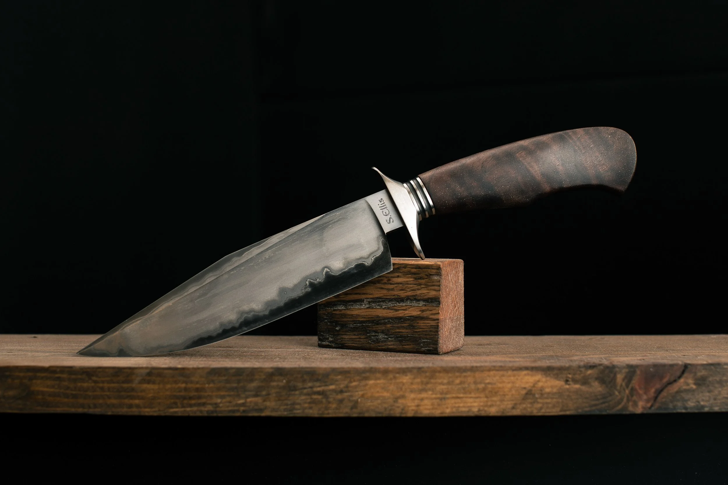 A high-quality kitchen knife with a marbled gray and black blade, dark wooden handle, and a metal bolster, balanced on a wooden block on a wooden surface against a black background.
