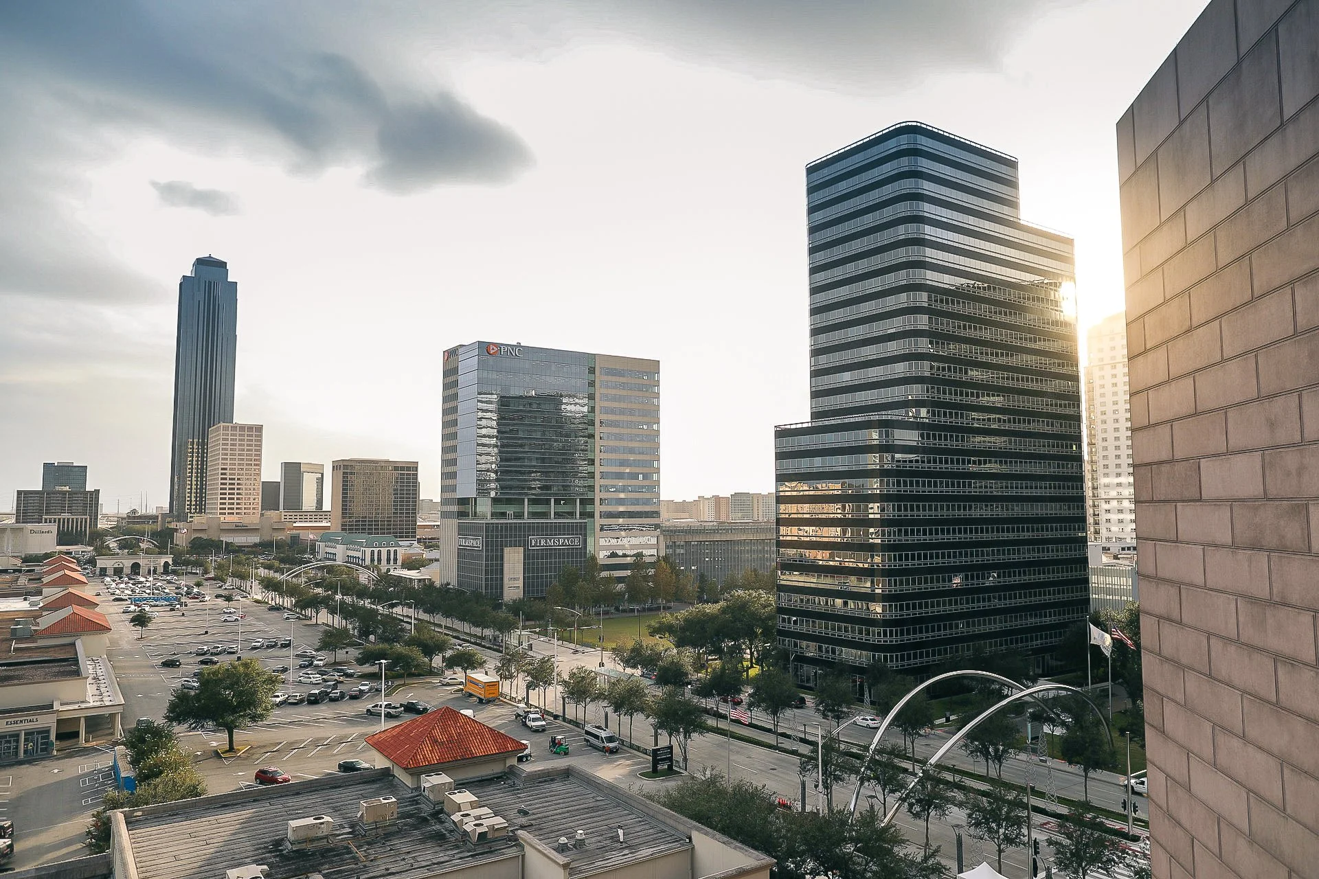 City skyline with tall modern office buildings, a parking lot, and a street with cars and trees, with the sun setting behind the buildings.