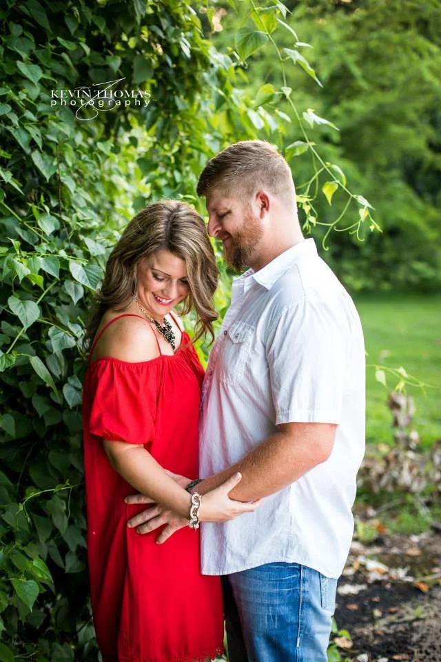 A young couple standing close together outdoors, smiling and looking into each other's eyes, with green leaves and bushes in the background.