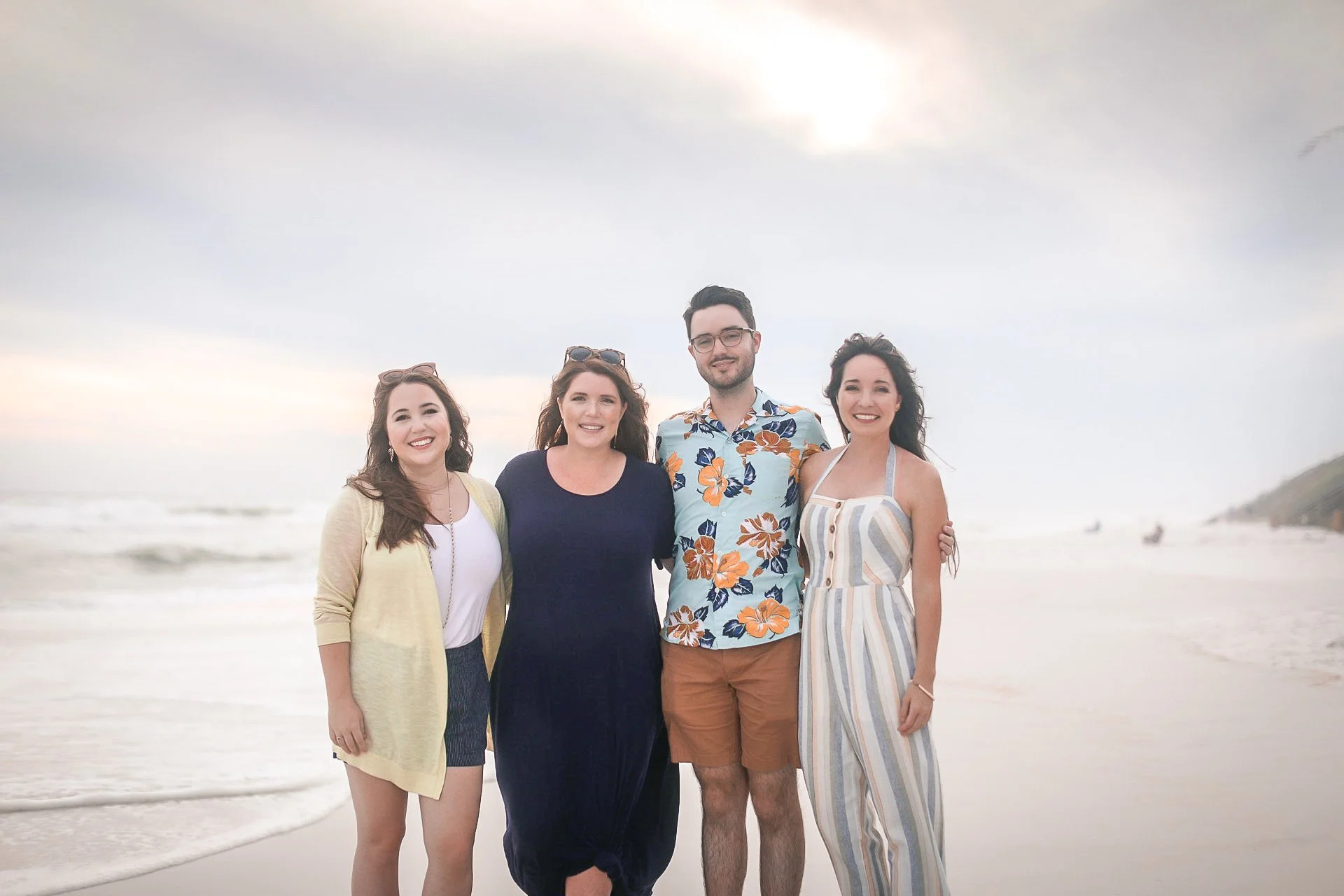 Four friends standing on the beach, smiling, with the ocean and cloudy sky in the background.