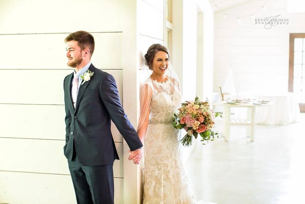 A bride and groom stand back-to-back indoors, holding hands. The bride is in a lace wedding dress and holding a bouquet, smiling. The groom is in a dark suit with a white boutonniere, looking to the side.