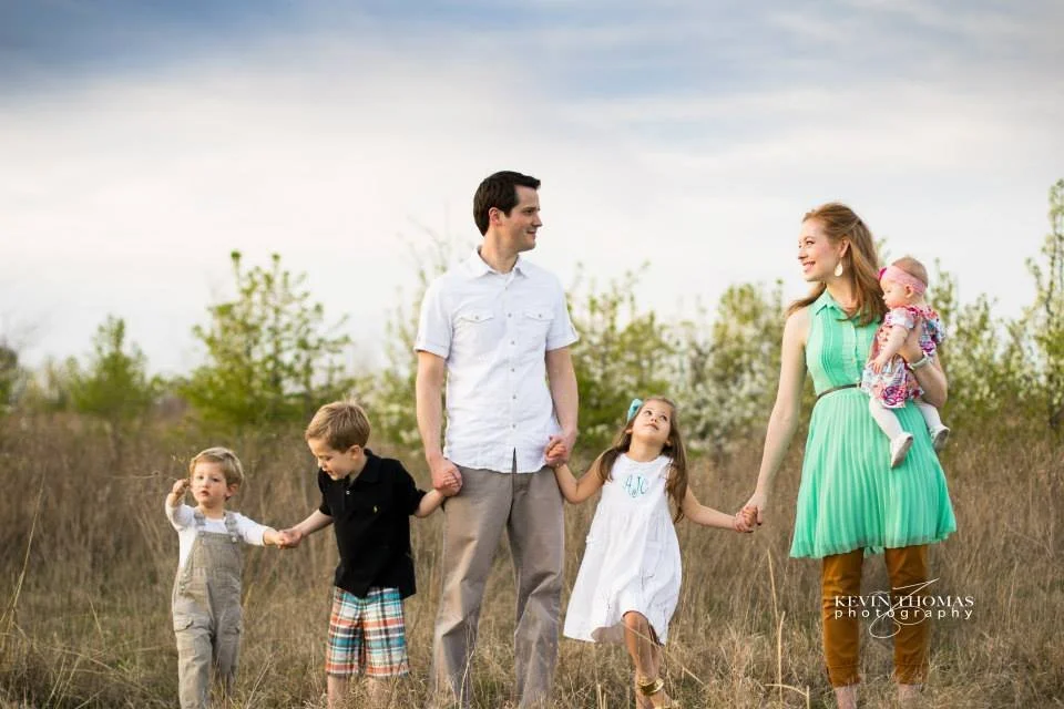 Family of six standing in a field holding hands, with trees and a cloudy sky in the background.