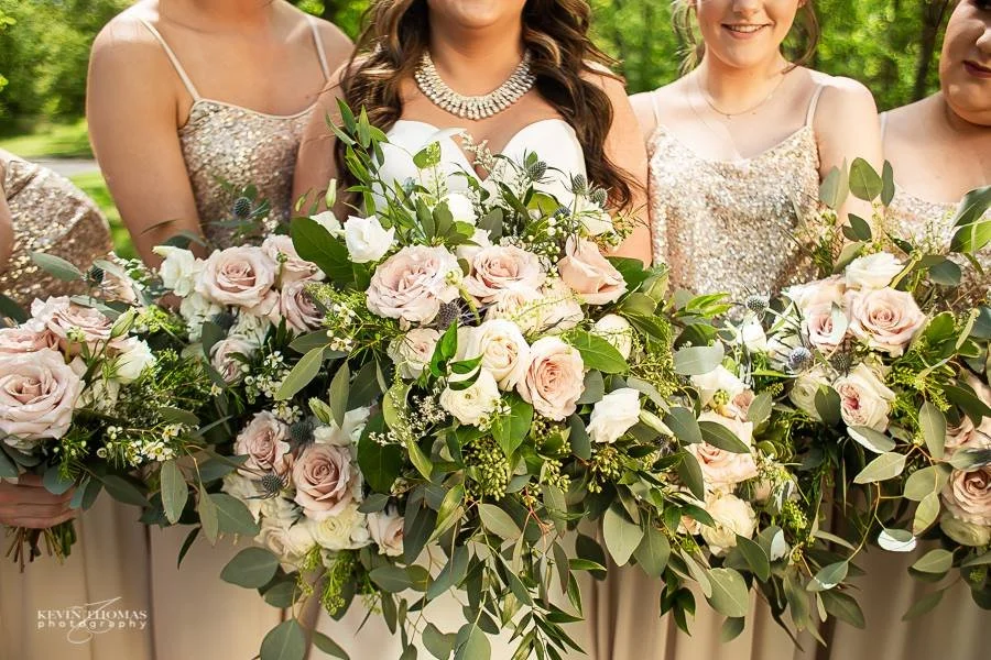 A bride and her bridesmaids holding bouquets with pink and white roses and greenery at a wedding.