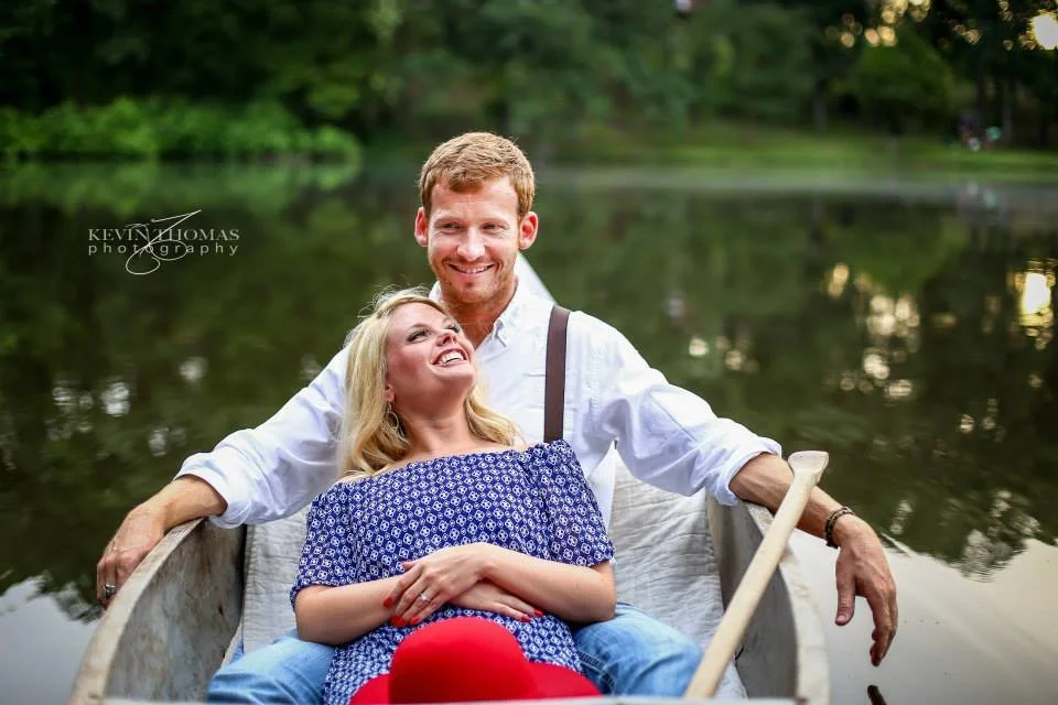 A couple smiling and enjoying a boat ride on a calm lake surrounded by trees.
