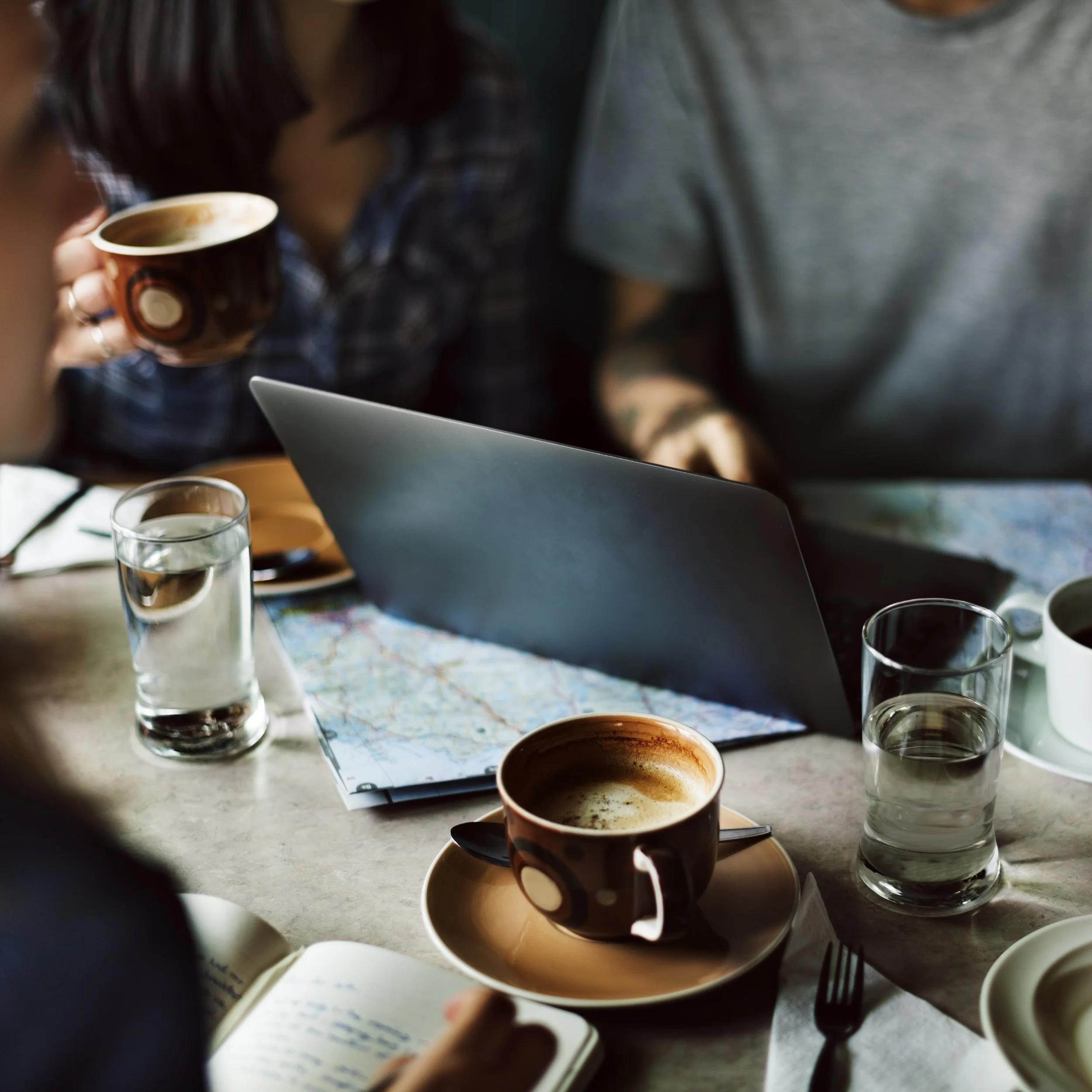 A group of people sitting at a table with coffee cups, glasses of water, a map, a laptop, and a notebook with handwritten notes.