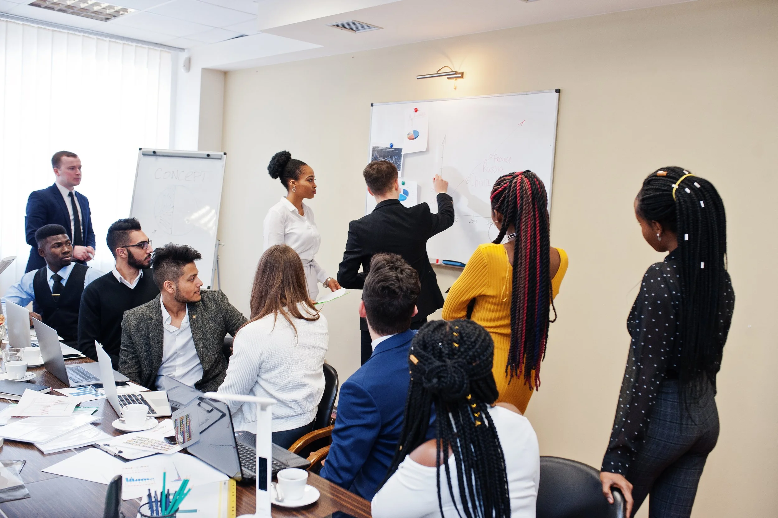 Business meeting with diverse professionals in a conference room, some writing on a whiteboard, others seated at a table with laptops and papers.