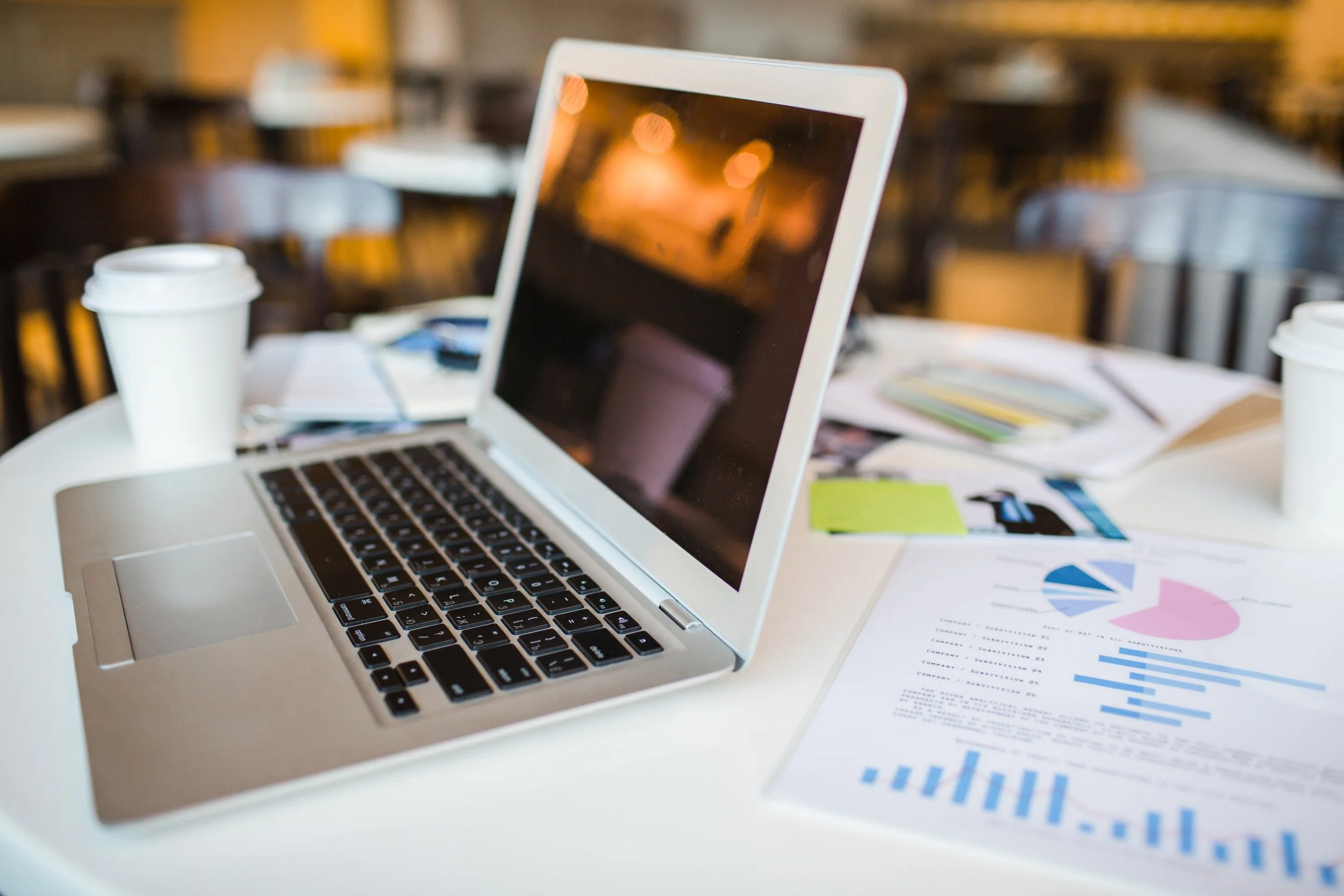 A laptop on a cluttered white table with sheets of paper containing charts and graphs, two disposable coffee cups, and blurred chairs in the background.
