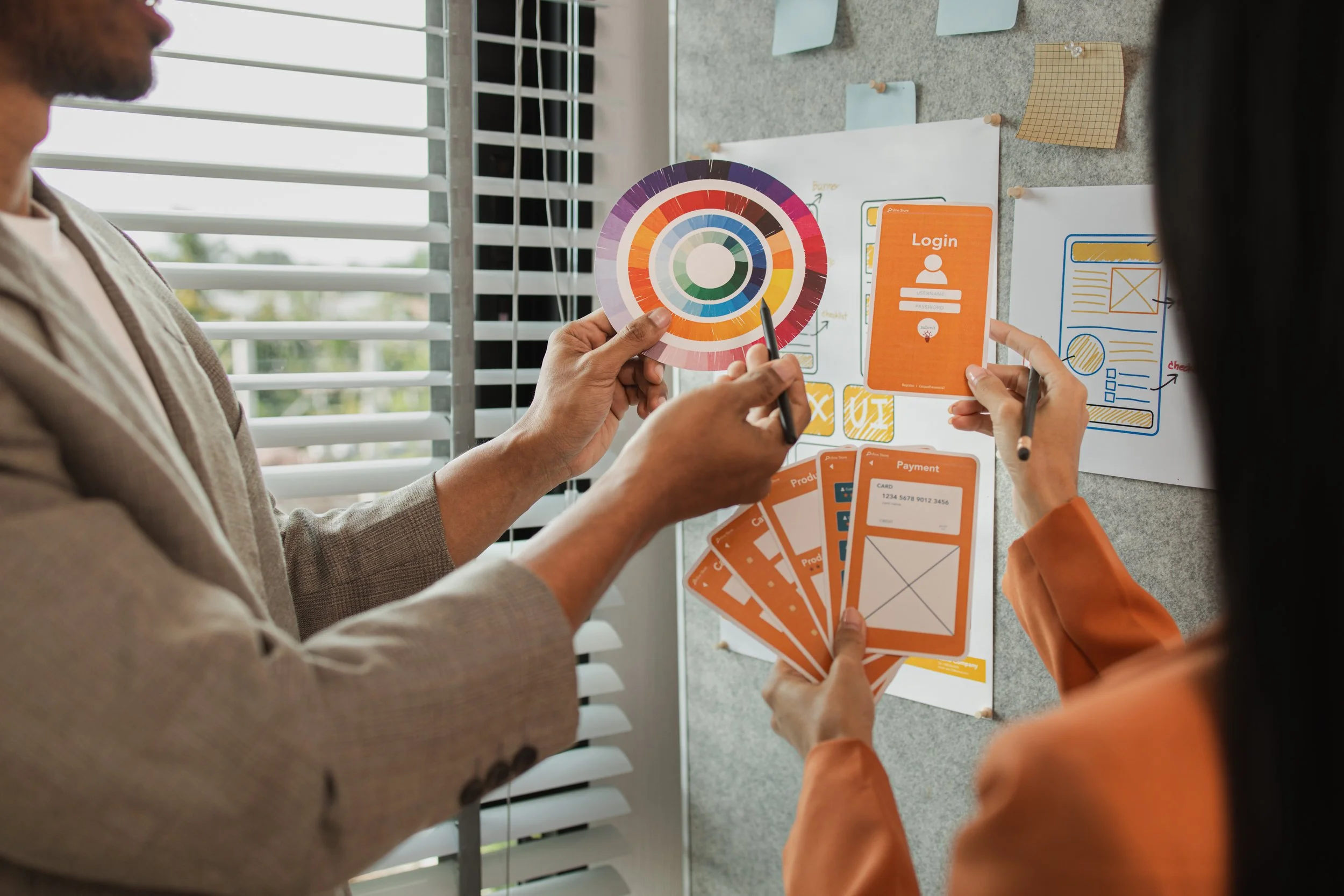 Two people working on a design project at a desk, holding color palettes and wireframes for a mobile app, with sketches and notes on the wall.