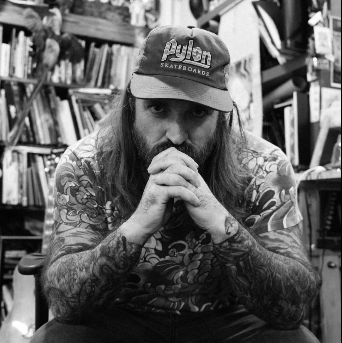 A man with tattoos on his arms, long hair, and a beard, sitting with his hands clasped in front of his face, wearing a cap that says 'Pylon Skateboards,' in a room with shelves filled with books and art supplies.