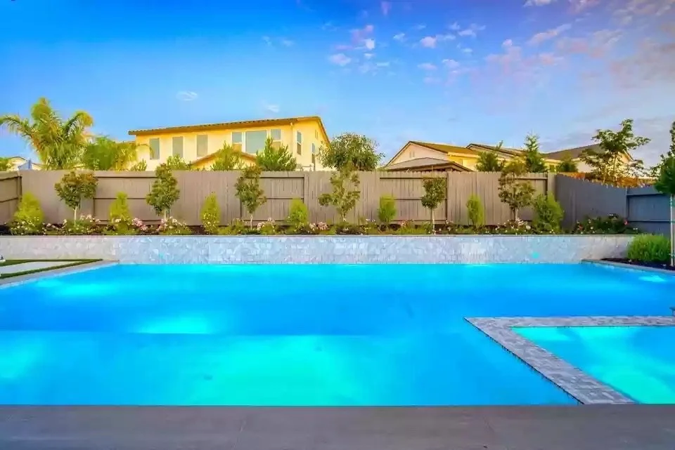 Backyard with a swimming pool, a wooden fence, trees, and a yellow house in the background, under a partly cloudy sky.