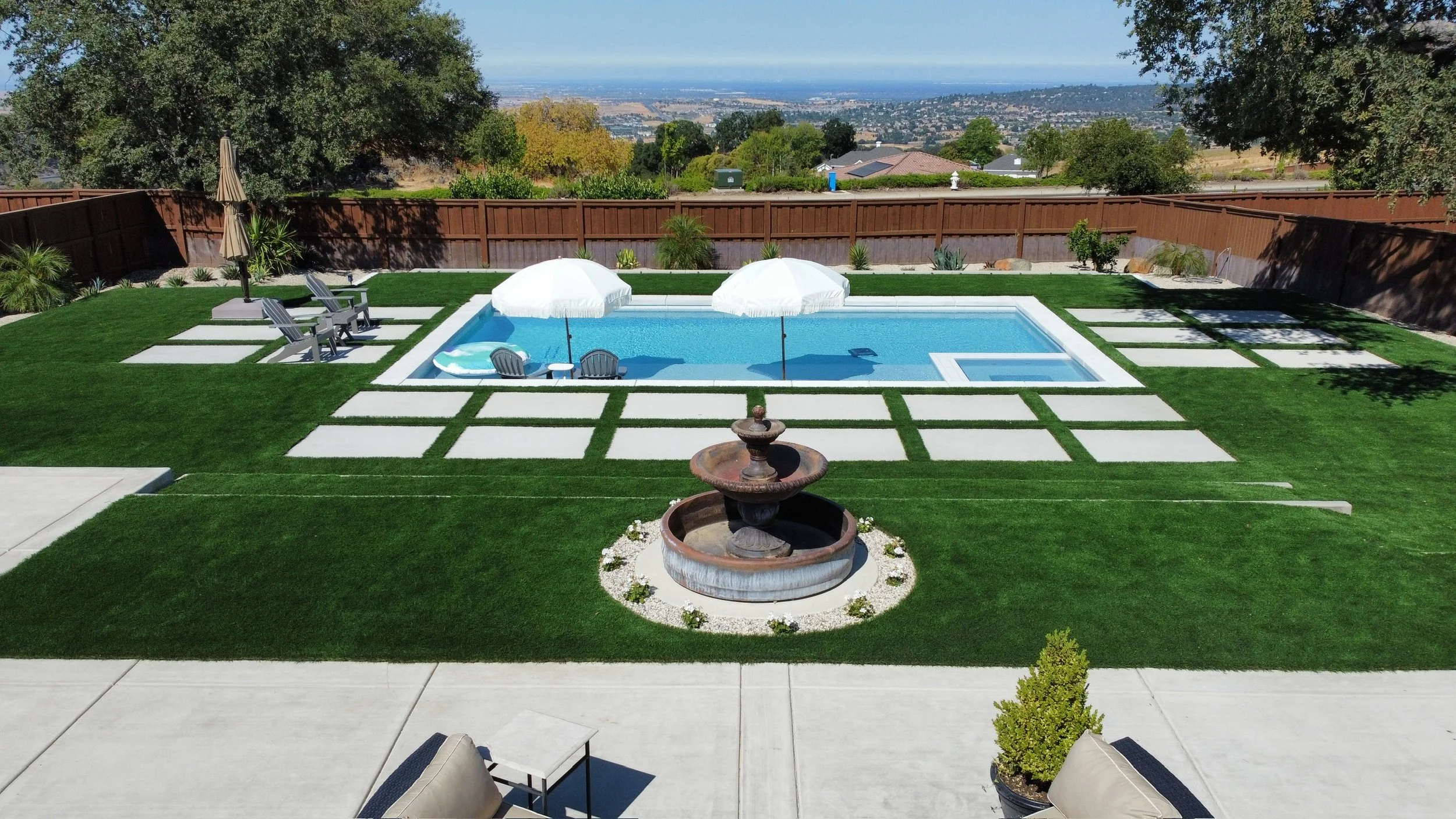 A backyard with a swimming pool, surrounded by green grass and decorative stone paths. There are two white umbrellas over the pool, with lounge chairs beneath them, set against a privacy fence. In the foreground, a fountain and outdoor seating are visible. The background shows a distant view of hills and a cityscape.