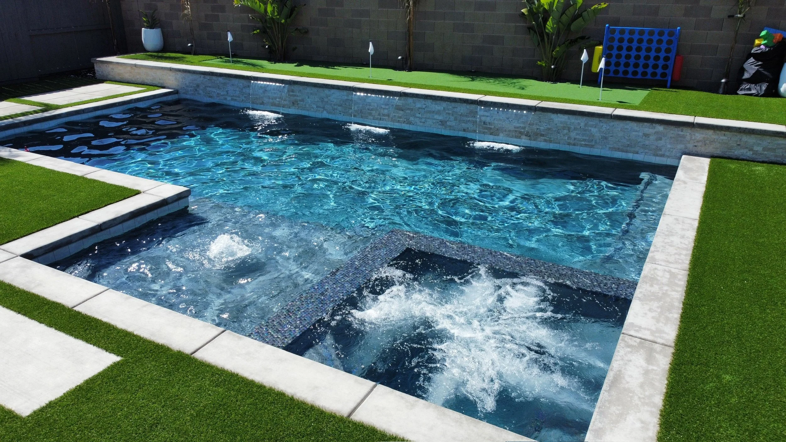 A backyard swimming pool with a built-in hot tub, surrounded by green artificial grass and concrete tiles. The pool features a water fountain wall along the back and a cornhole game on the grassy area - landscaping design done by Combat Pools