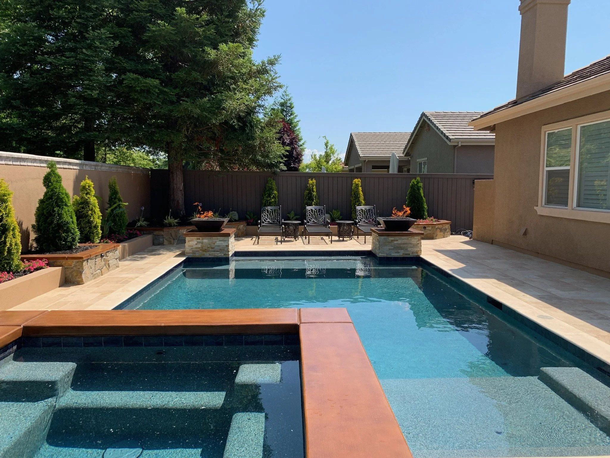 Backyard with a swimming pool, surrounded by a stone patio, with four lounge chairs and small tables, a large tree, and a fence with plants along it. A house with tan walls and a window is visible on the right.
