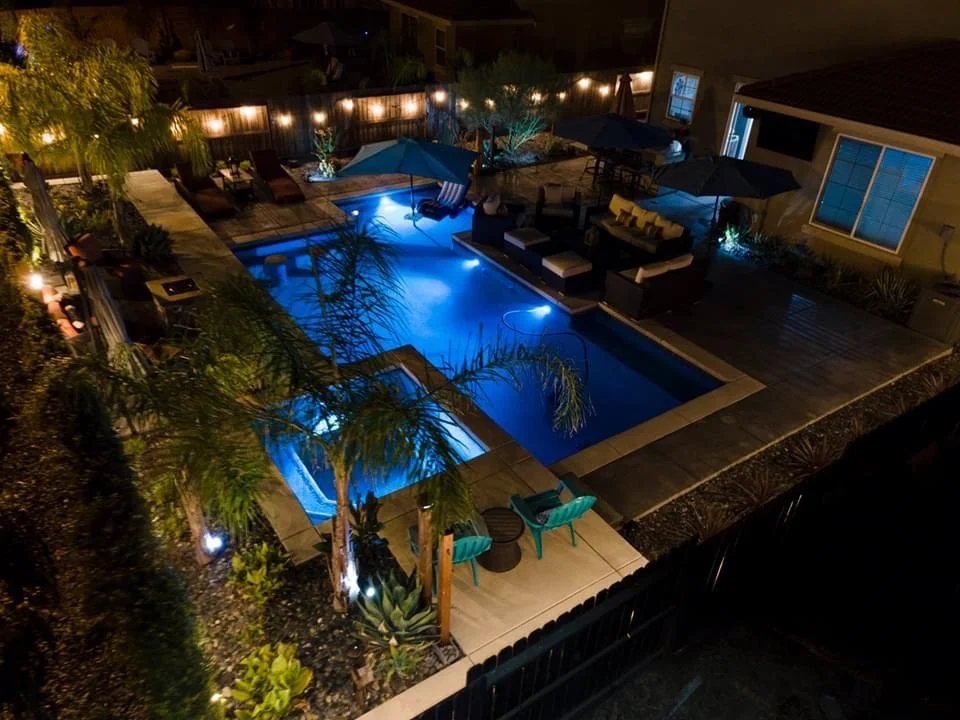 Night view of a backyard swimming pool illuminated with blue lights, surrounded by outdoor furniture, umbrellas, trees, and string lights along the fence, with a house on the right side.