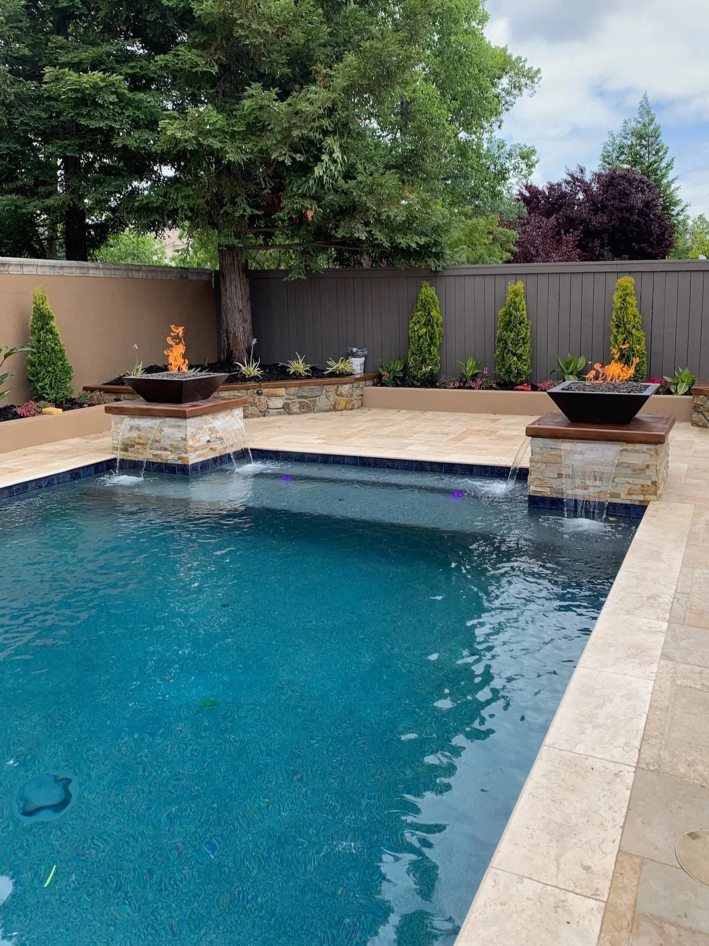 A backyard swimming pool with two decorative fire features on stone pedestals at each end, surrounded by a tan tile deck, with landscaped greenery and trees in the background.