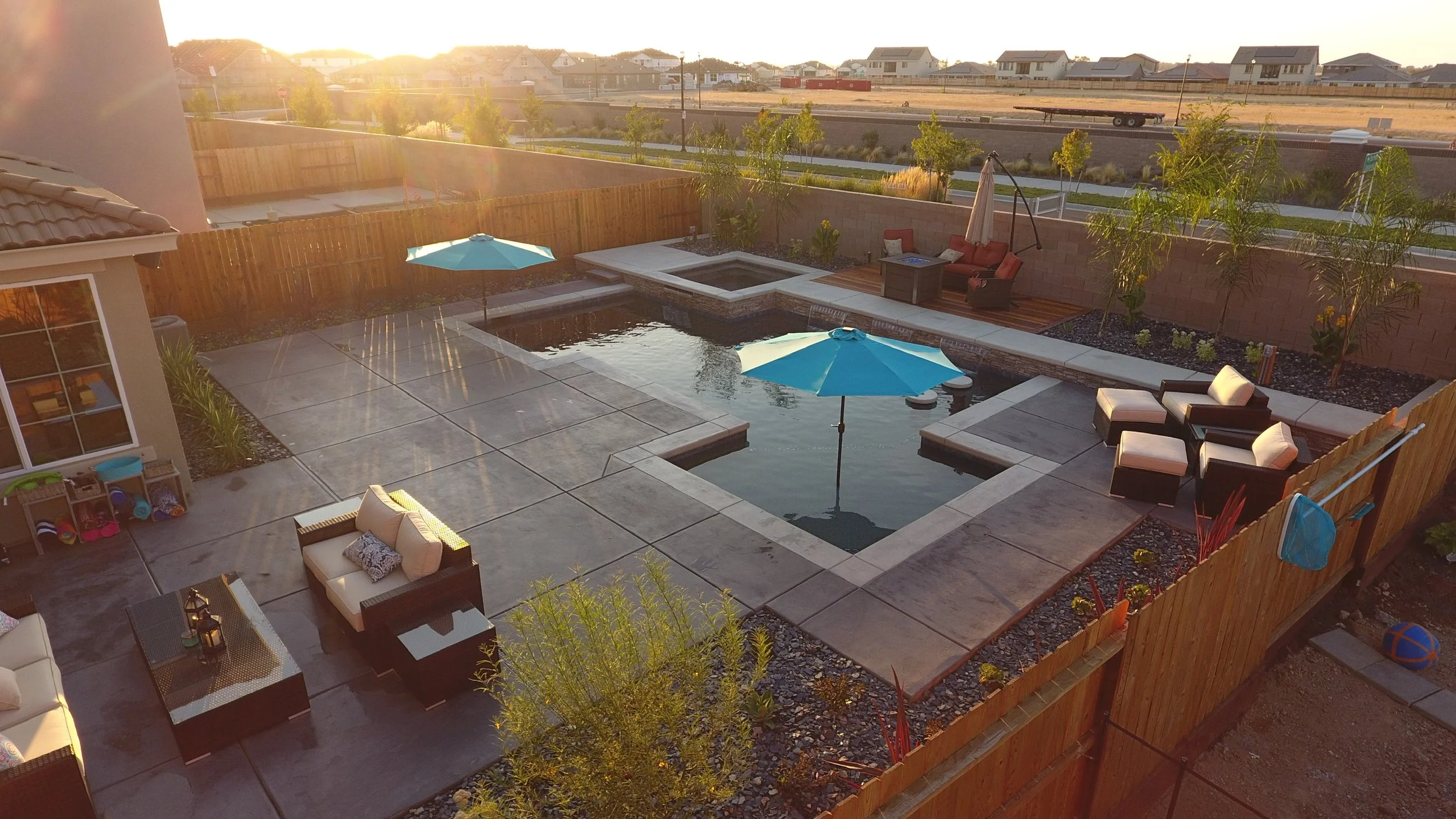 A backyard with a swimming pool, outdoor seating, umbrellas, and a hot tub, enclosed by a wooden fence, during sunset.