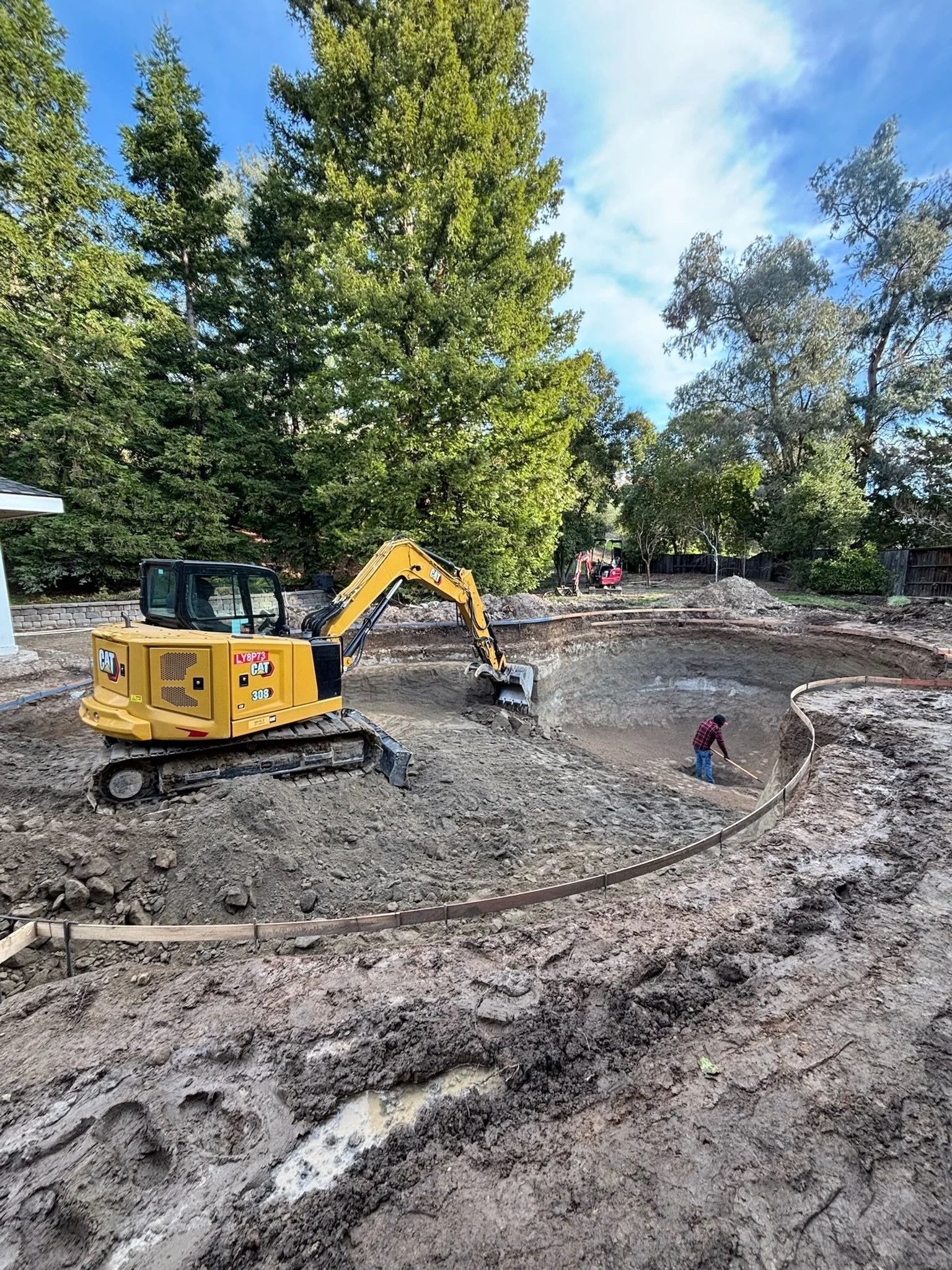 Construction site with excavator and worker digging in the ground for a swimming pool, surrounded by trees and clear sky.