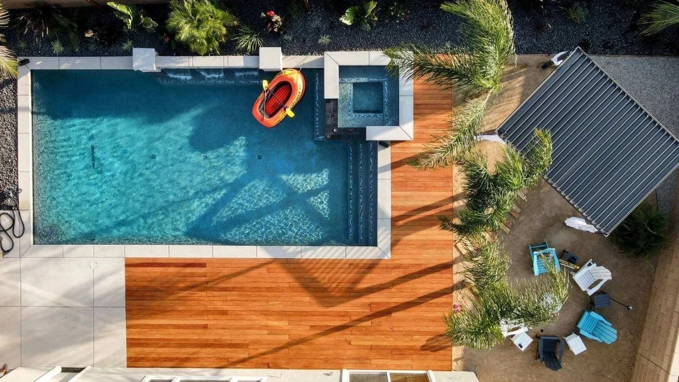 Top-down view of a backyard with a swimming pool, wooden deck, palm trees, outdoor furniture, and a small covered area.