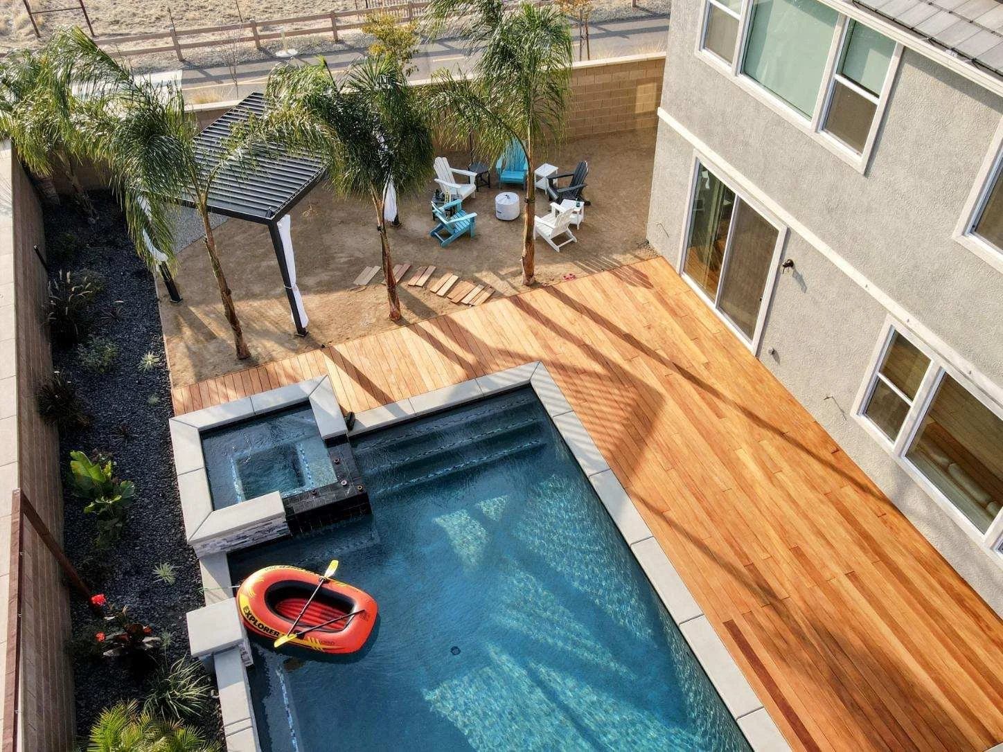 A backyard with a swimming pool, a wooden deck, and a small area with palm trees, outdoor chairs, a gazebo, and a kayak in the pool.