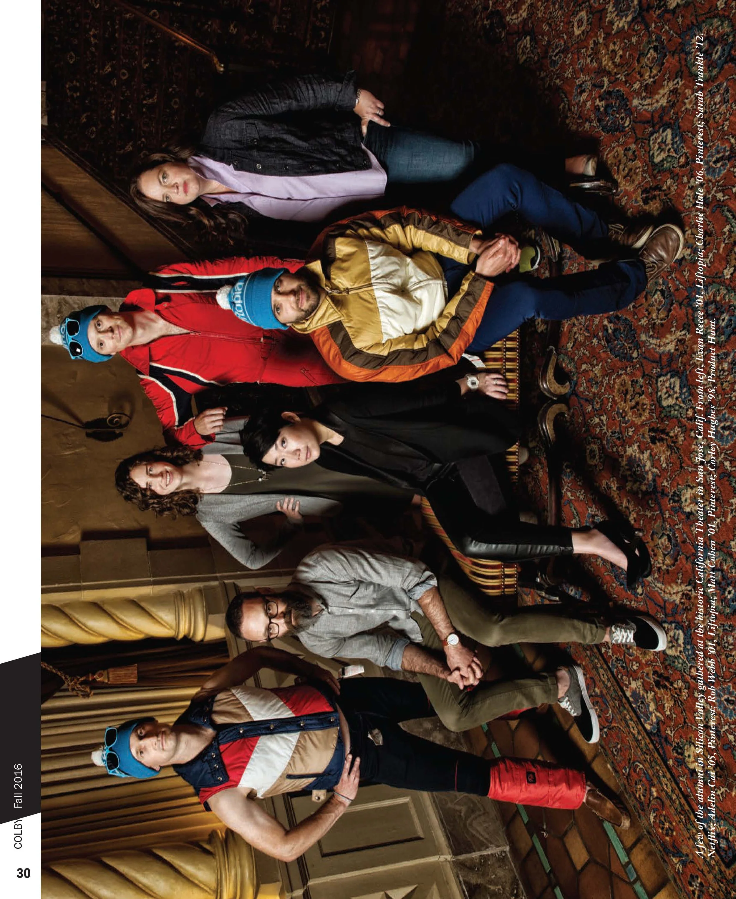 Group of six people, five women and one man, posing together in a room with ornate decor and a patterned carpet. Some are standing, while others are sitting on furniture.
