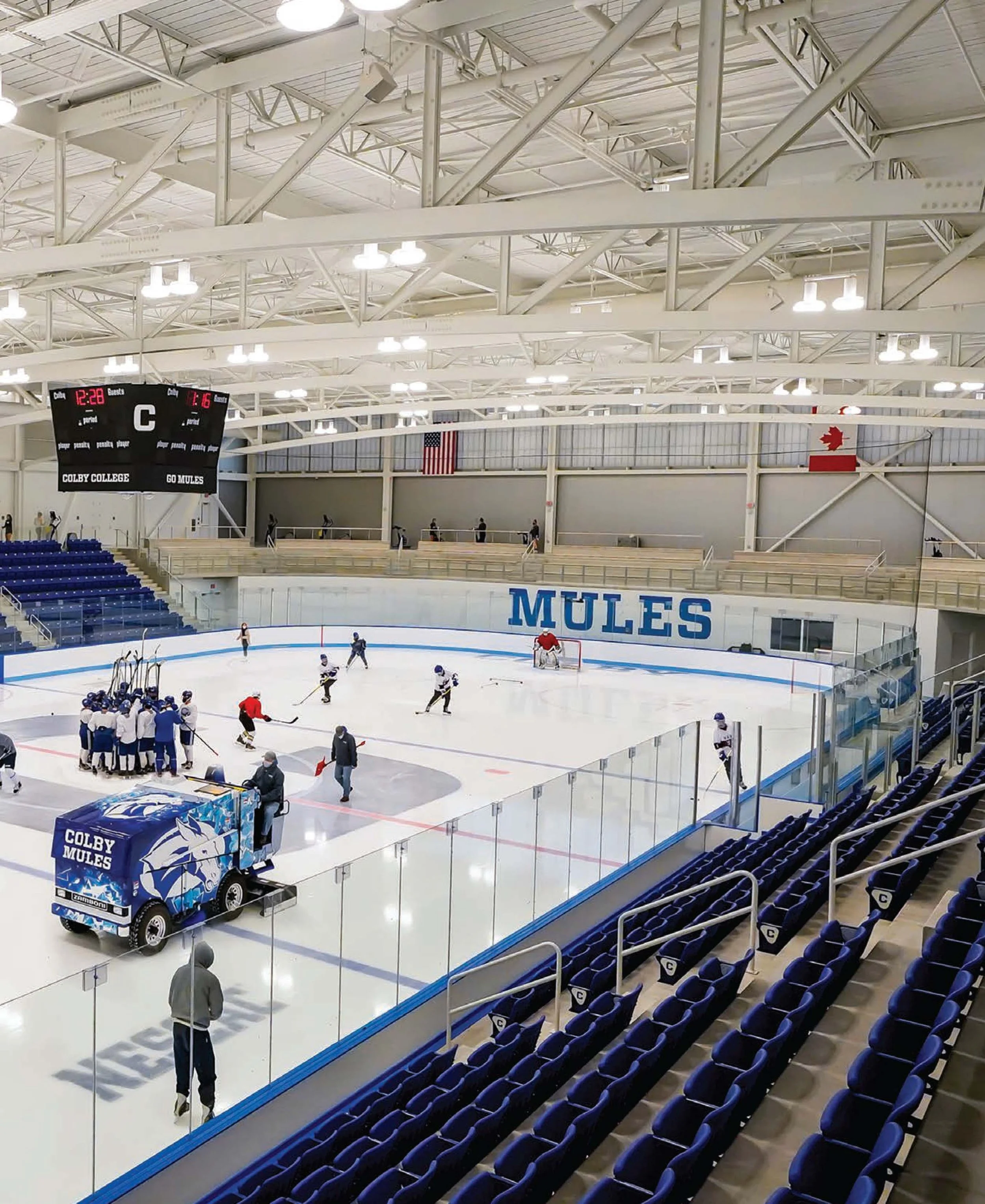 Inside an ice hockey rink with players warming up on the ice, a scoreboard showing 12:28 and 6 minutes, blue seats on the sides, and the word "MULES" on the wall.