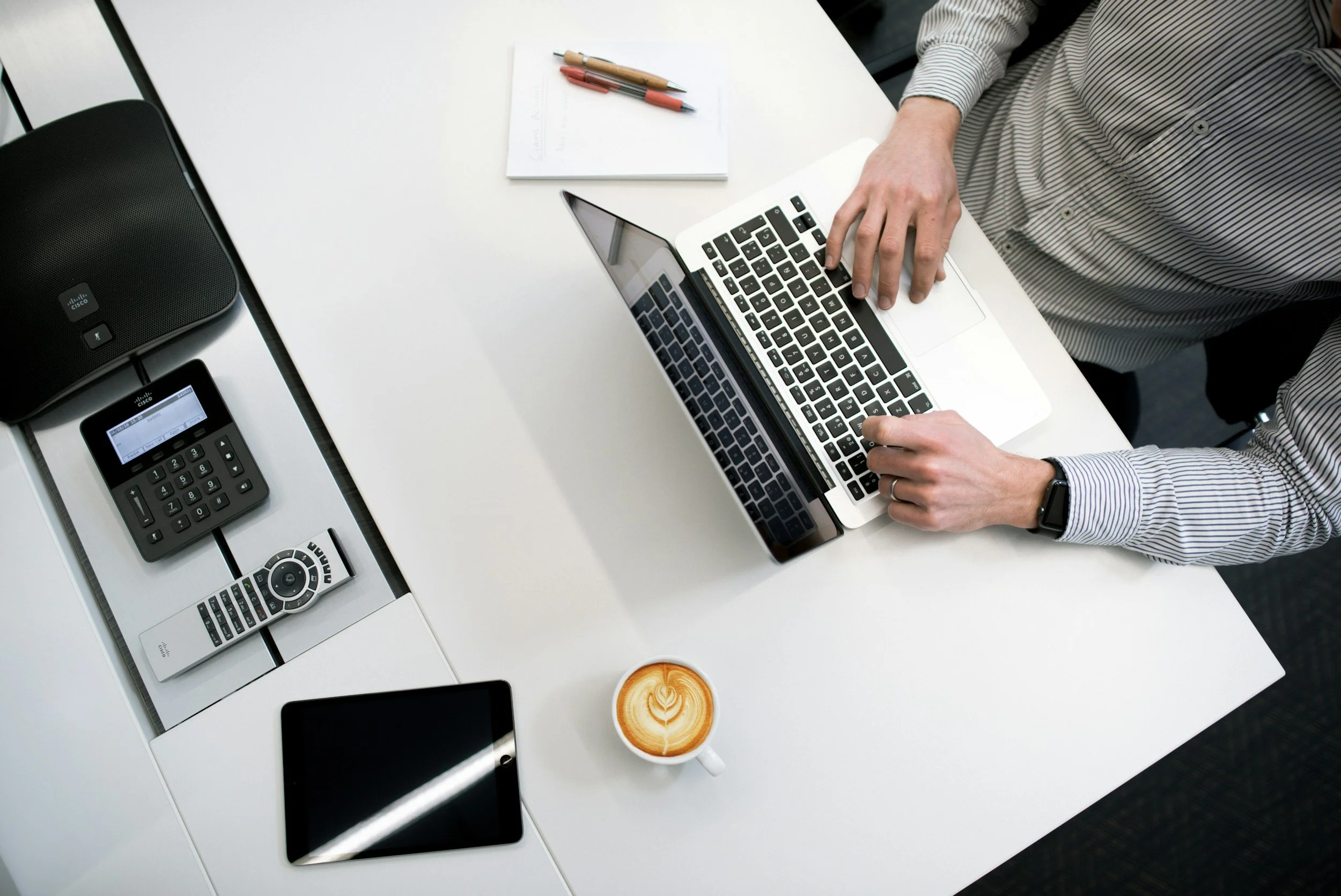 A person in a striped shirt working on a laptop at a white desk, with a coffee cup, tablet, remote control, a notebook with pens, a conference phone, and a smartphone on the desk.