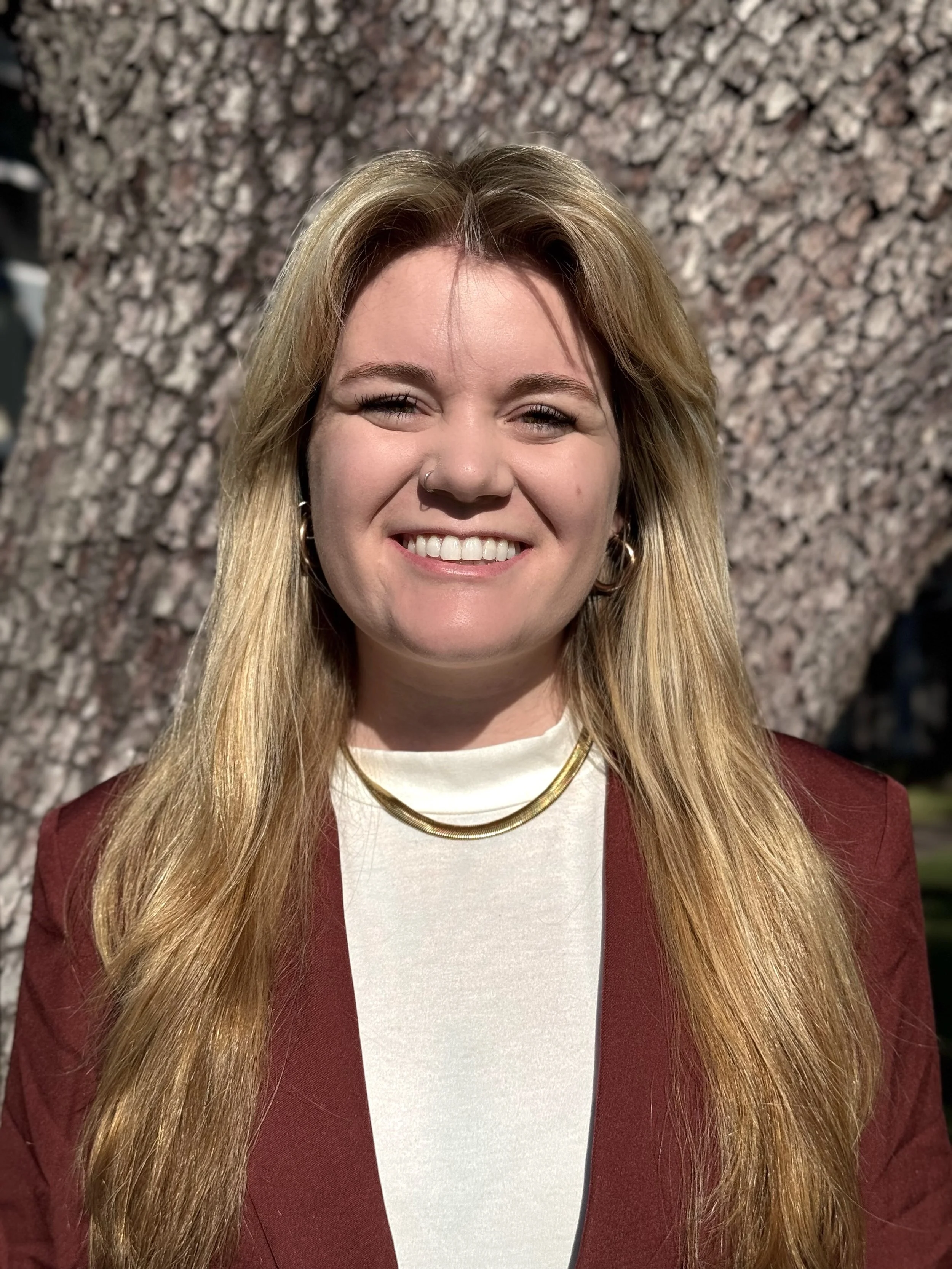 A woman with long blonde hair smiling in front of a textured brown and pink granite wall, wearing a white top, a red blazer, and gold jewelry.