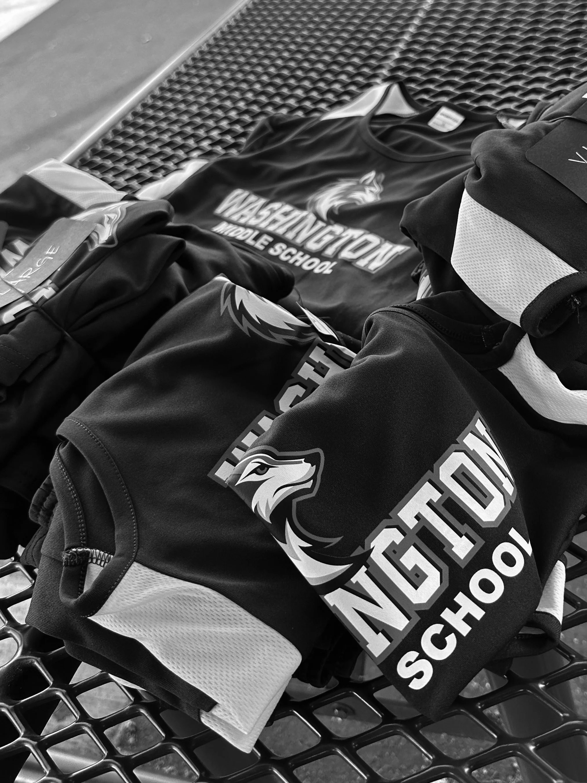 A black and white photo of a shopping cart with a black T-shirt, a black and white hoodie, and a black bag inside. The T-shirt has a logo and text for Washington Middle School, featuring an eagle.