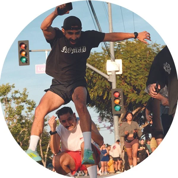 Two men joyfully playing on a city street during daylight, with traffic lights and pedestrians in the background. One man, wearing a black t-shirt and black shorts, is jumping on the other's back, who is squatting and wearing a white t-shirt and red shorts.