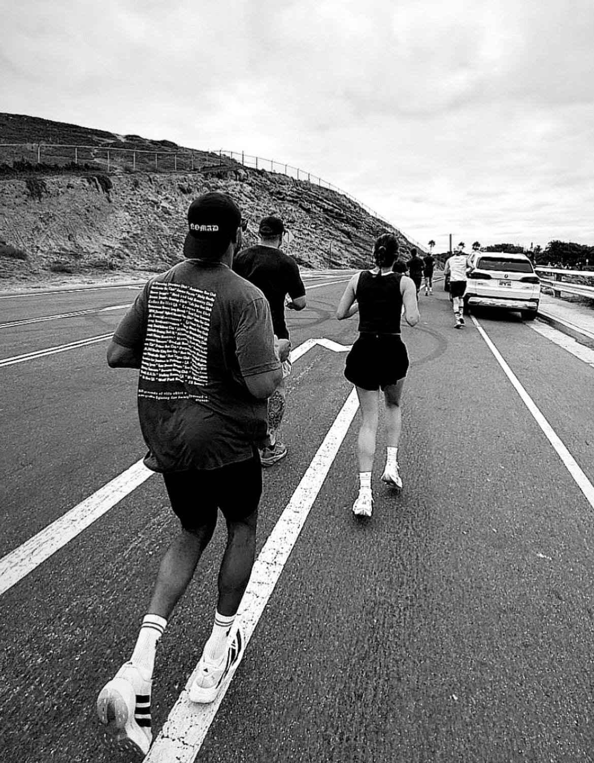 A group of runners, including a woman in a black tank top and shorts, running along a road with a parked car on the side, hillside in the background, and cloudy sky.