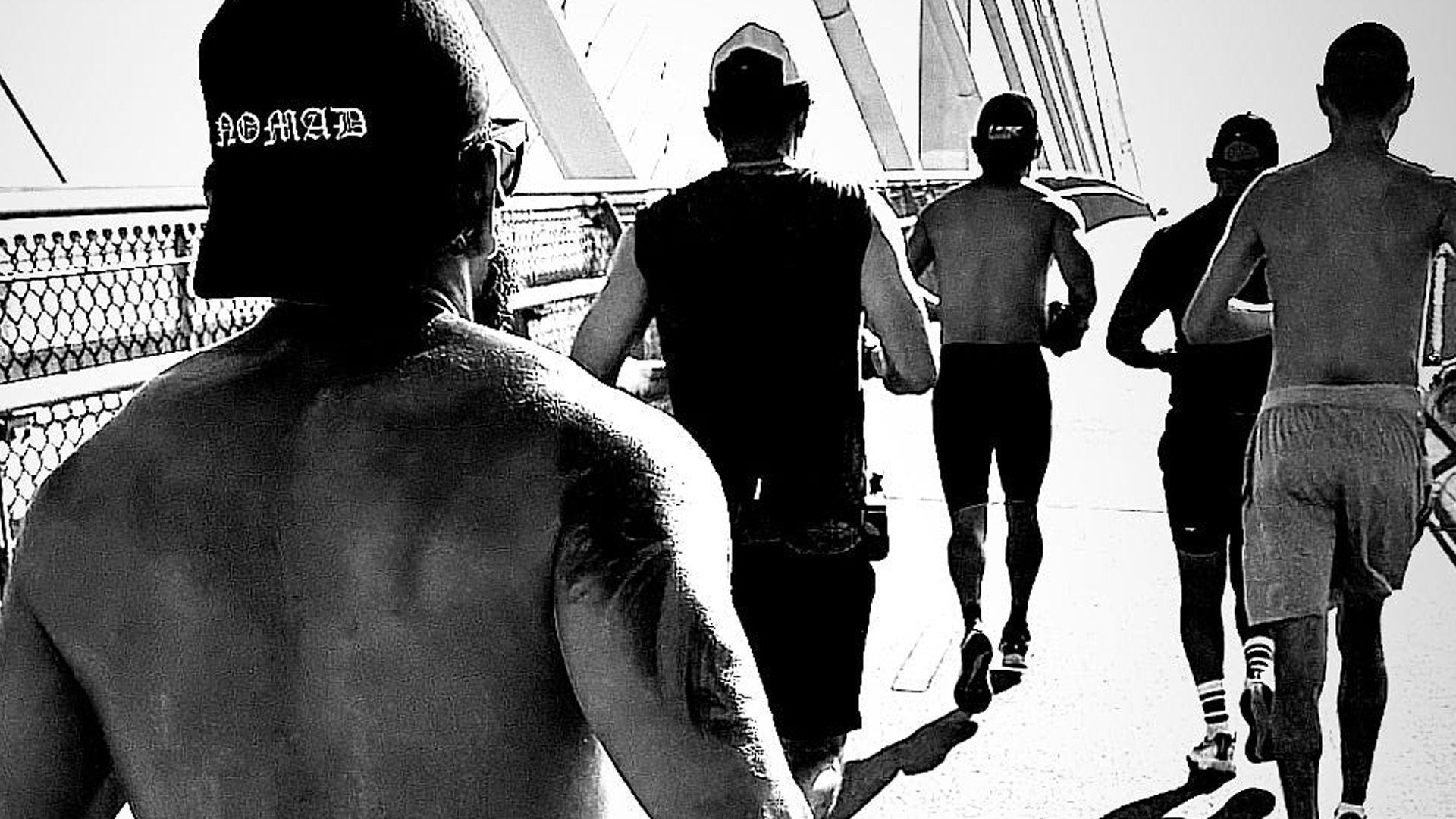 A black and white photo of several runners, some shirtless and some wearing shirts or tank tops, running on a bridge.