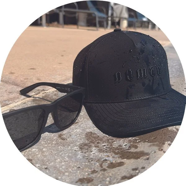 A black baseball cap with the word 'damn' embroidered on it, and a pair of black sunglasses, both placed on a concrete surface outdoors.