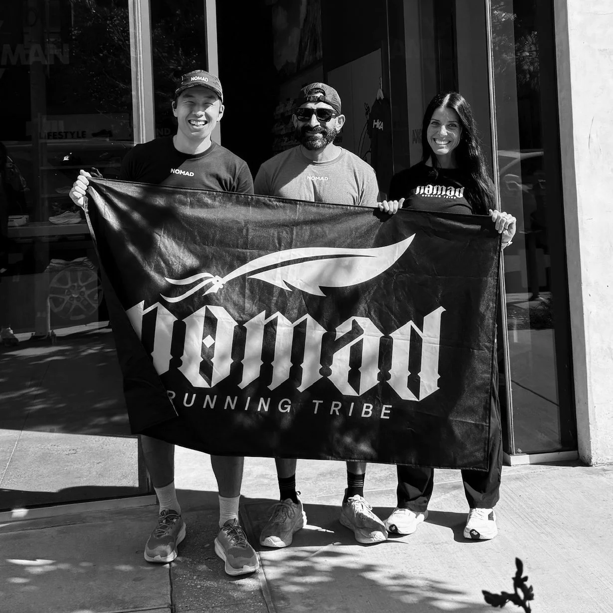 Three people standing outside a building, holding a flag with the words "Nomad" and "Running Tribe" and a feather logo, smiling at the camera.