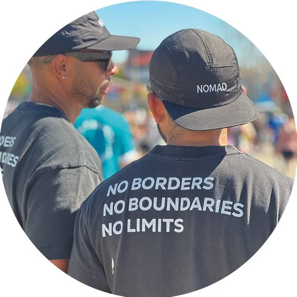 Two men wearing dark shirts and hats, with one shirt reading 'NO BORDERS NO BOUNDARIES NO LIMITS,' standing outdoors with a crowd of people in the background.