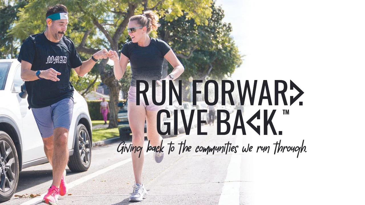 A man and woman running together on a sunny street, engaging in a high-five or fist bump, with trees and cars in the background. The text on the image reads 'Run Forward. Give Back.' with a subtitle 'Giving back to the communities we run through.'