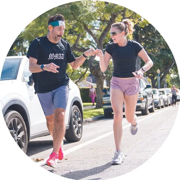 A man and woman running together on the street, smiling and giving each other a high-five, with parked cars and trees in the background.