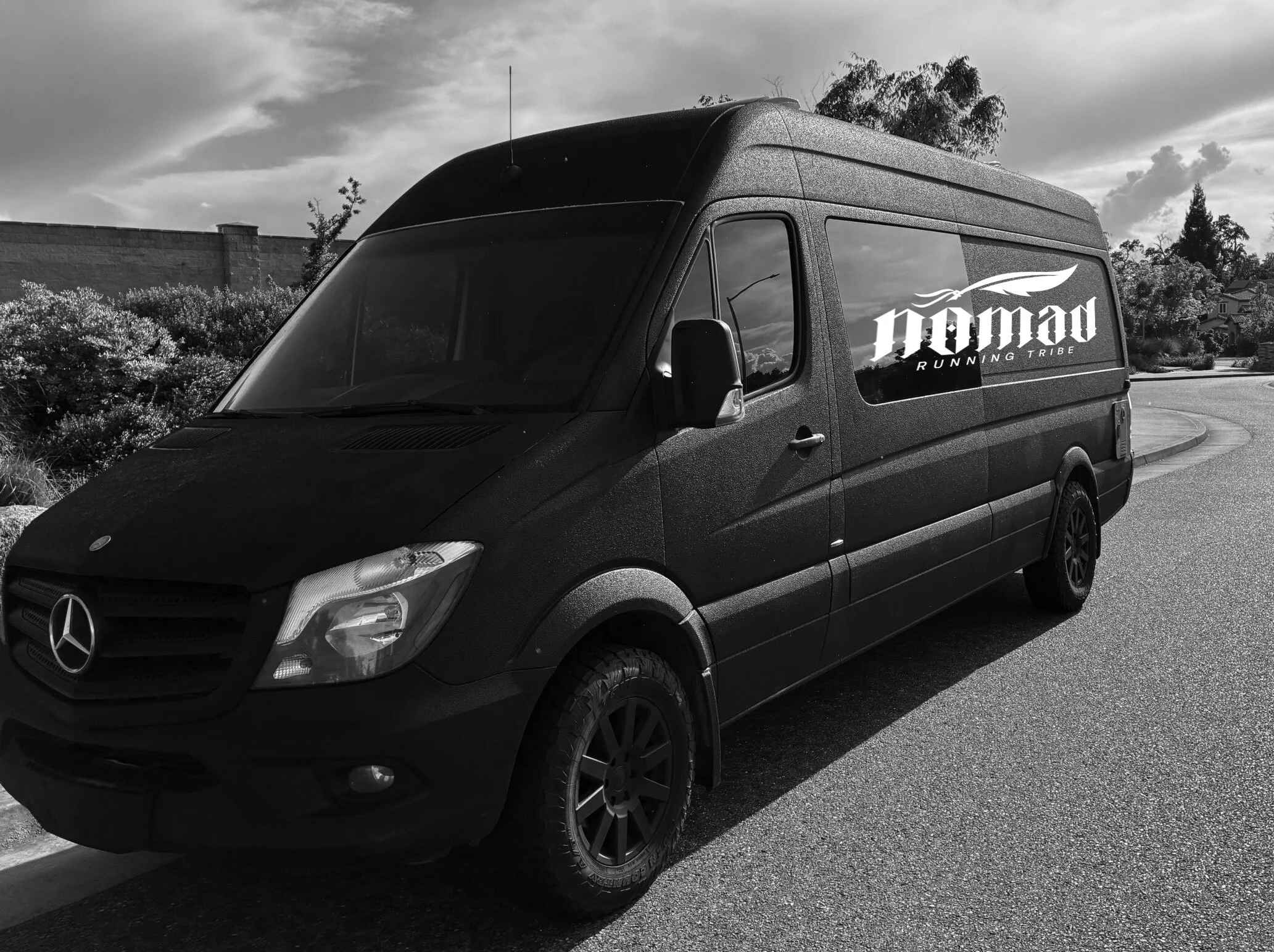 A black Mercedes-Benz van parked on a street with a large sticker on the side that reads 'nomad' and 'Running Tribe' with a feather logo, during cloudy weather.