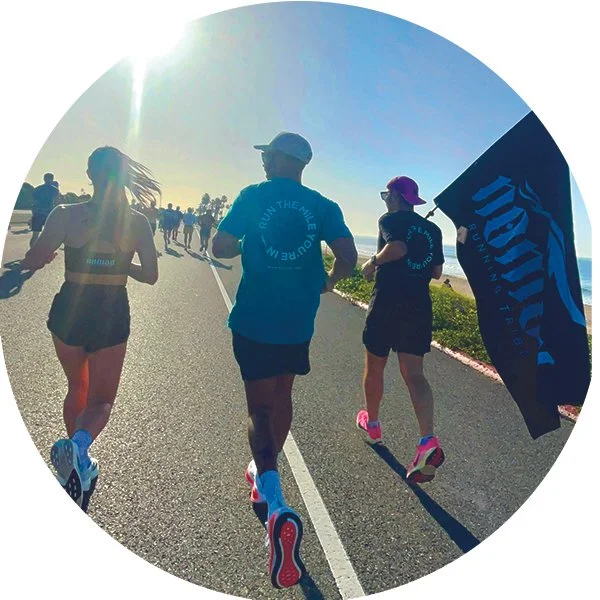 Group of runners participating in a marathon along a scenic coastal road with the sun shining brightly overhead, one woman holding a flag.