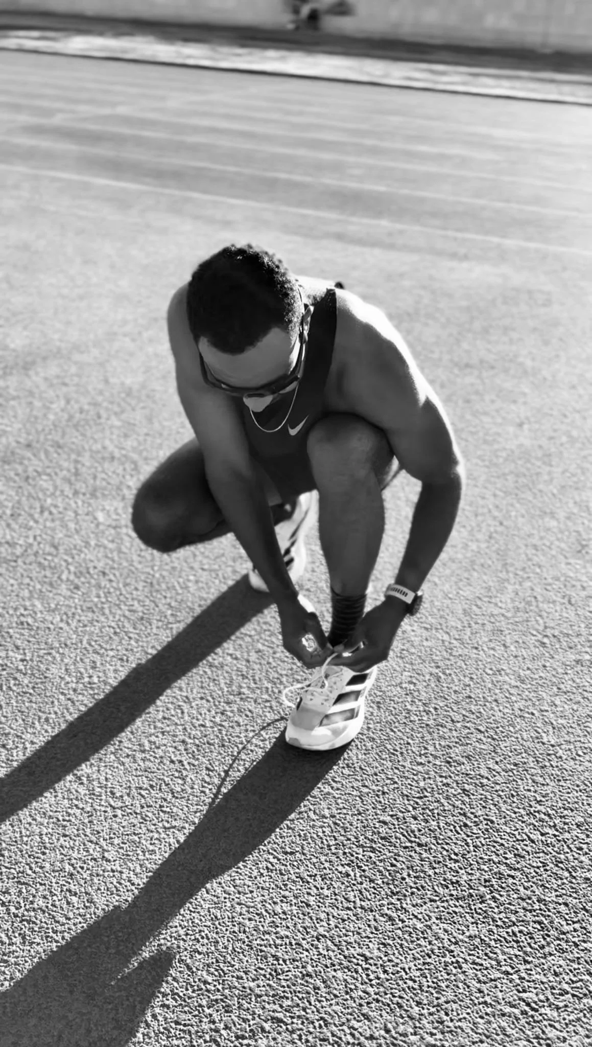 A male athlete in running gear, including sunglasses and a waist pack, is crouched down on a running track tying his shoelaces under bright sunlight, casting a long shadow.