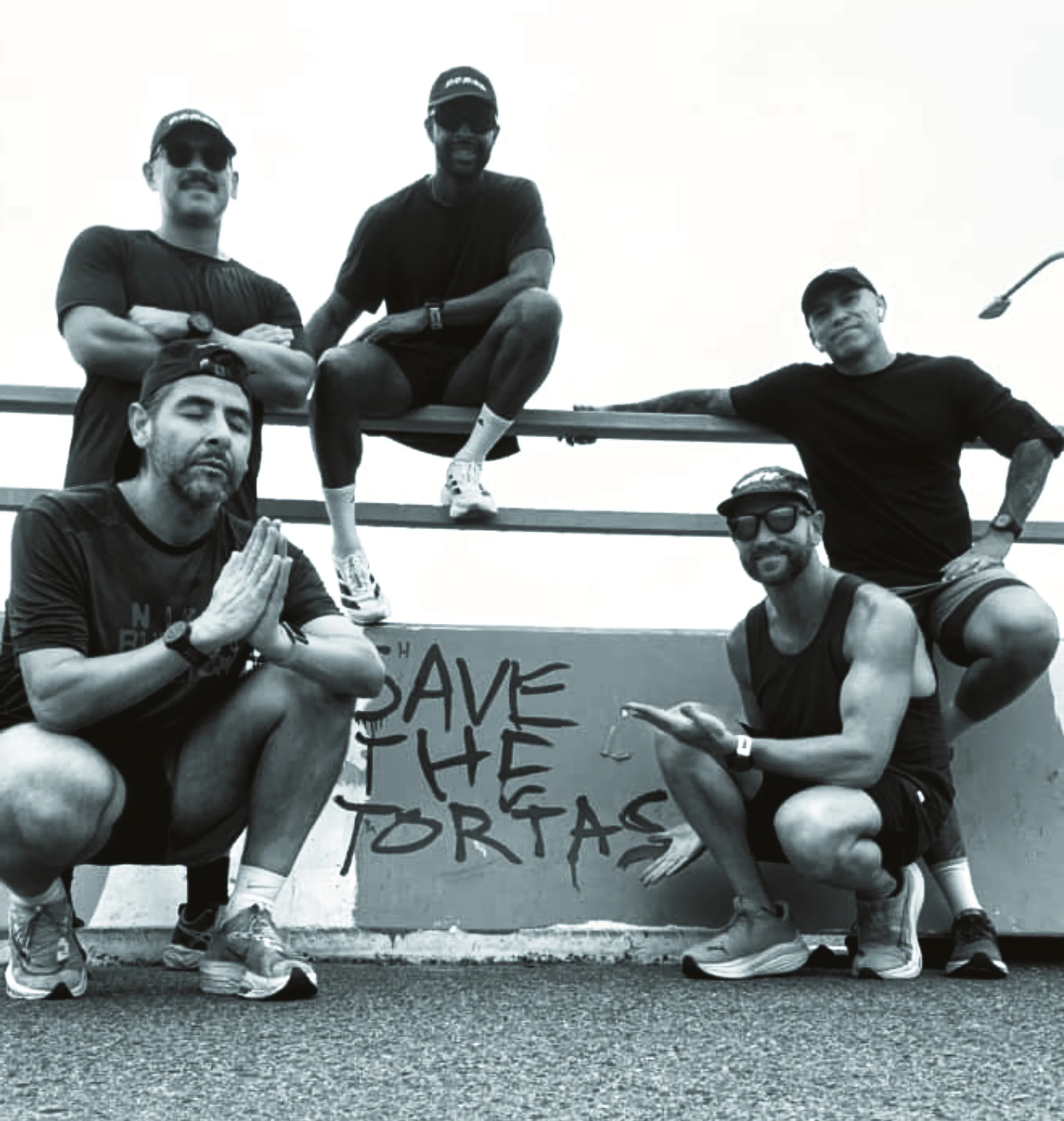 Six people posing outdoors near a graffiti-covered structure that says "Save the Torta". Four are crouching, two are sitting on top of the structure. All are wearing casual athletic clothing and sunglasses.