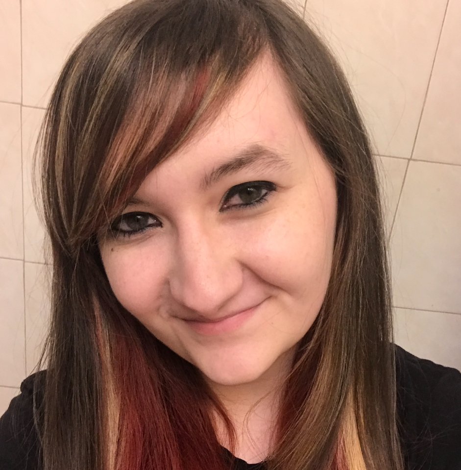 Close-up of a smiling woman with brown hair and red highlights, in front of a tiled wall.