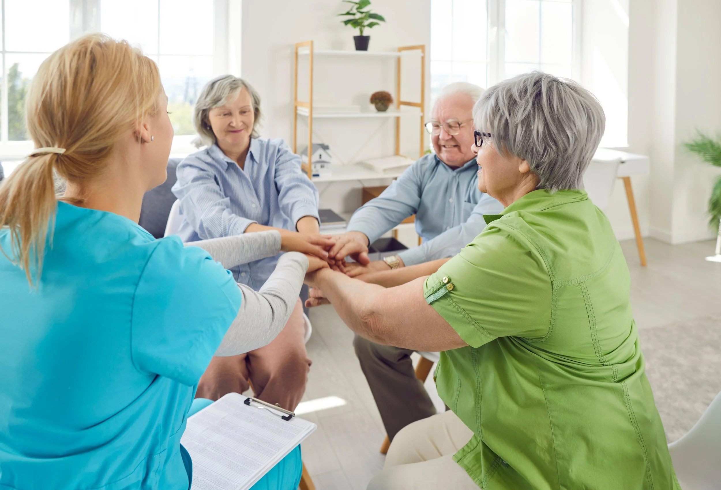 Group of elderly people and a caregiver holding hands in a circle during a support group meeting in a bright, modern room.