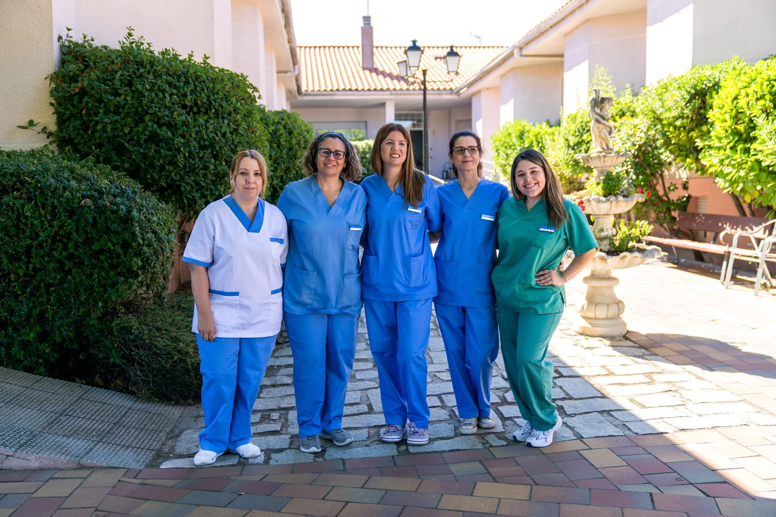 Five healthcare workers standing outdoors on a cobblestone path in front of green bushes, a decorative fountain, and a building with tiled roof, all smiling at the camera.