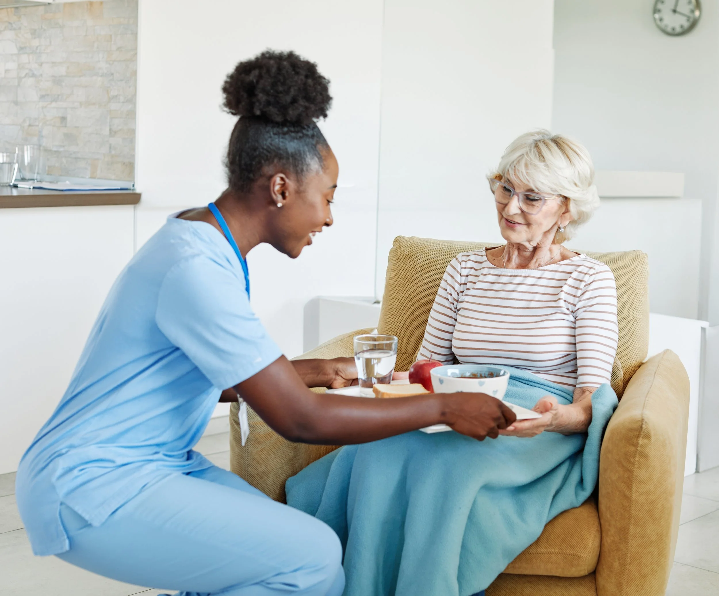 A nurse serving a meal to an elderly woman sitting in a recliner chair in her home.