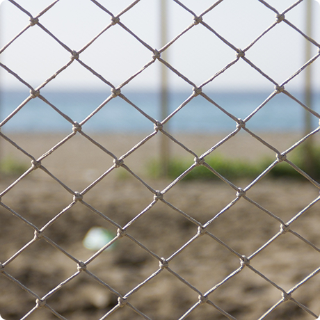 View of a soccer ball on a dirt field seen through a chain-link fence with a body of water in the background.