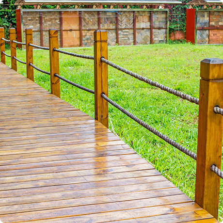 Wooden boardwalk with a wooden post and rope fence, next to a grassy area and brick wall in the background.