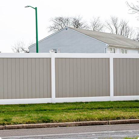 A beige backyard fence with a white top, a grassy lawn, and a street curb in front of it. A house and leafless trees are in the background.