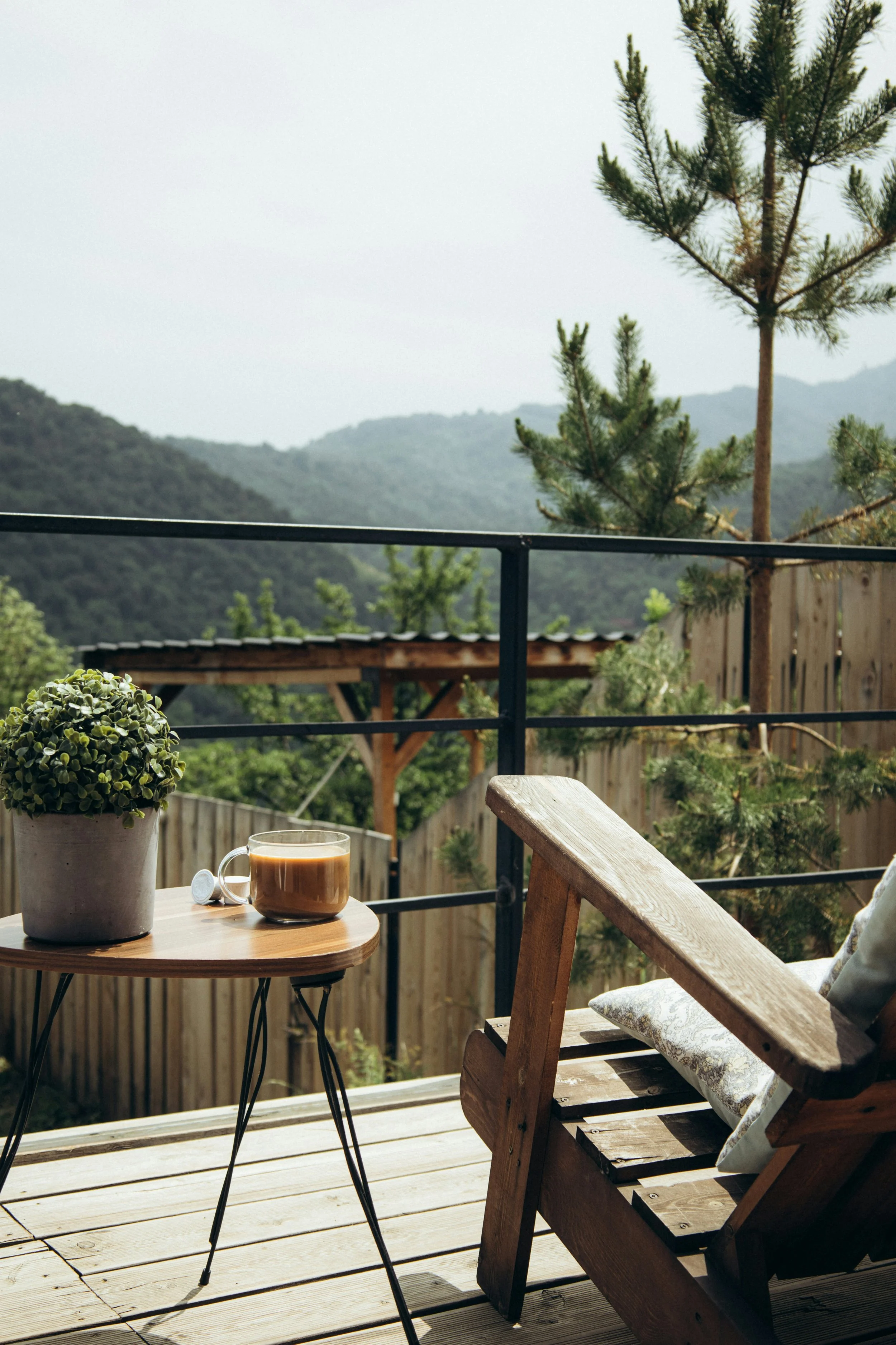 A cozy outdoor wooden deck with a small round table holding a potted plant, a coffee cup, and a spoon. A wooden chair with a floral cushion is nearby. In the background, there is a view of green hills, trees, a wooden fence, and a cloudy sky.